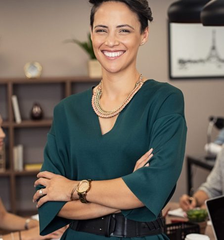 Portrait of successful business woman standing with her colleagues working in background at office. Portrait of cheerful fashion girl in green dress standing with folded arms and looking at camera. Beautiful businesswoman feeling proud and smiling.